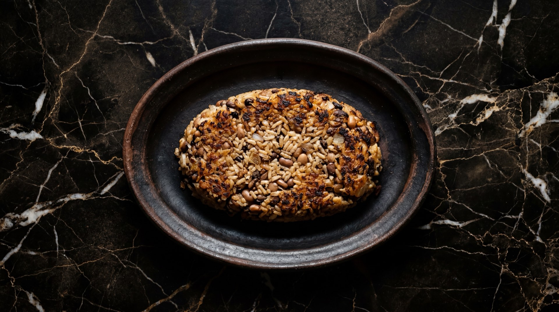 Tacu tacu peruano dorado y crujiente de arroz blanco cocido con frejoles canario sobre plato de ceramica negra mate en mesa de marmol negro con vetas doradas