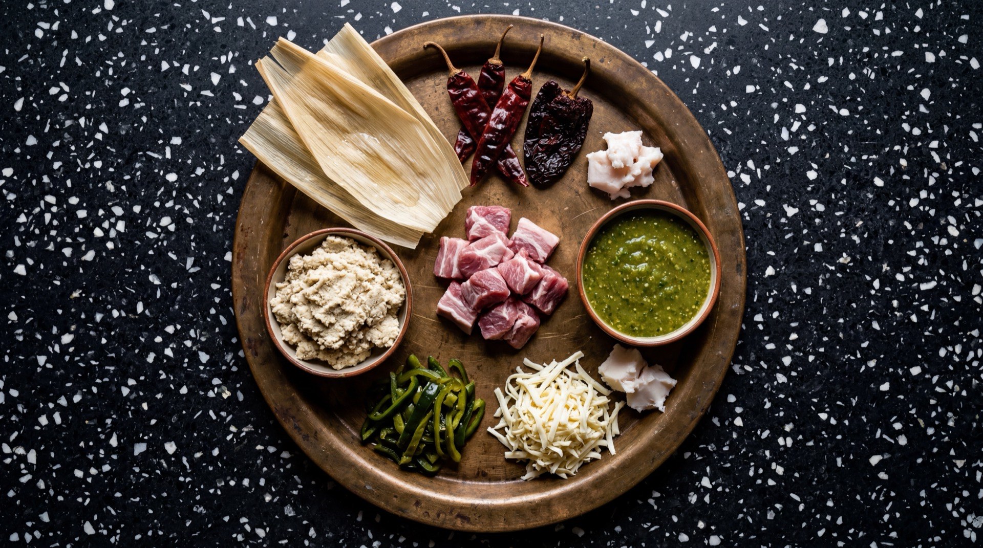 Ingredientes para tamales mexicanos: masa de maiz para tamales nixtamalizada, manteca de cerdo a temperatura ambiente, hojas de maiz secas remojadas, pechuga de pollo cocida y deshebrada, tomates verdes pelados, chiles serranos frescos, cebolla blanca mediana, dientes de ajo pelados, cilantro fresco lavado y sal de mesa