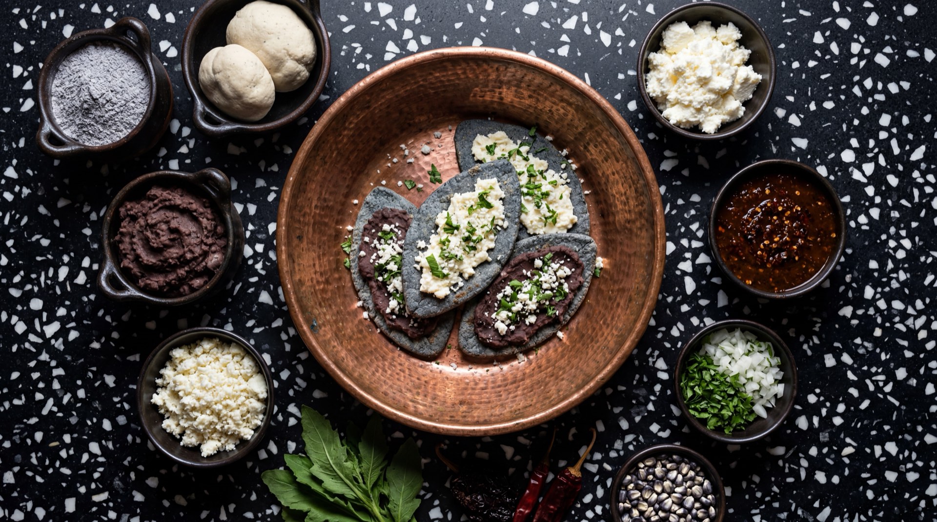 Ingredientes de tlacoyos mexicanos: masa de maiz azul nixtamalizado fresca, frijol negro refrito espeso, nopales tiernos cortados en cubitos, queso fresco desmoronado, salsa verde de tomate de milpa, cebolla blanca finamente picada, crema agria mexicana y cilantro fresco