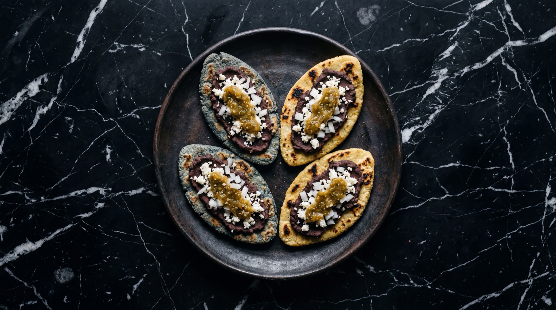 Tlacoyos mexicanos de maiz azul con nopales asados y queso fresco desmoronado sobre plato de piedra volcanica en mesa de granito negro con vetas doradas
