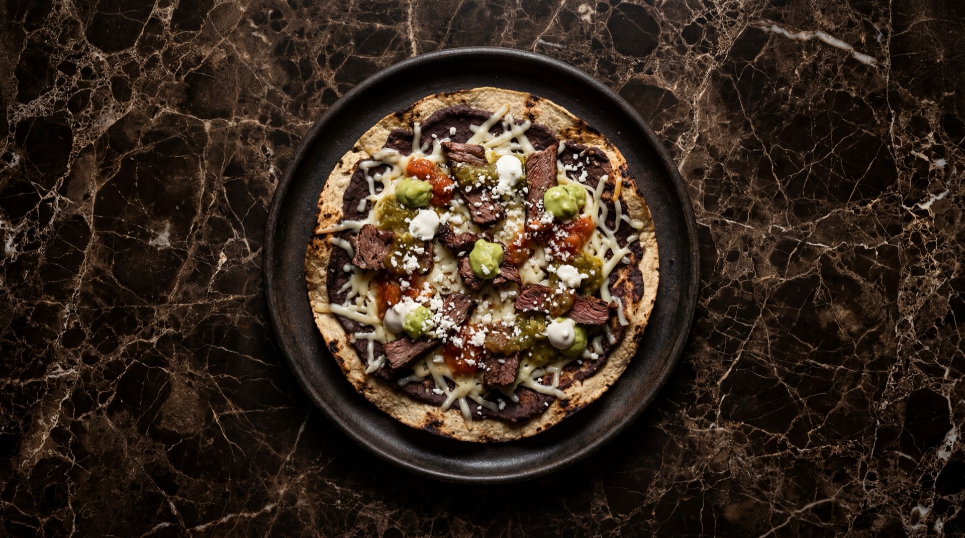 Tlayuda oaxaquena de tasajo de res con queso Oaxaca, frijoles negros y aguacate en comal de barro sobre mesa de piedra volcanica oscura