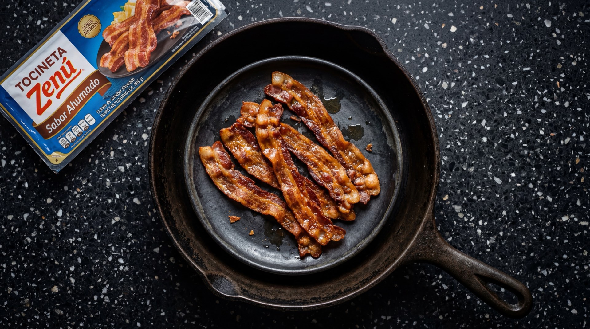 Tocineta Zenu ahumada en tajadas crocantes doradas sobre sarten de hierro fundido negro con fondo de marmol oscuro