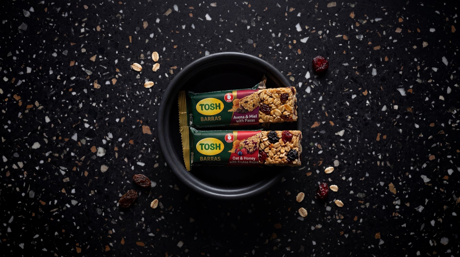 Barras Tosh de Noel en empaque verde individual sobre mesa de granito oscuro, barra de granola con avena y miel visible