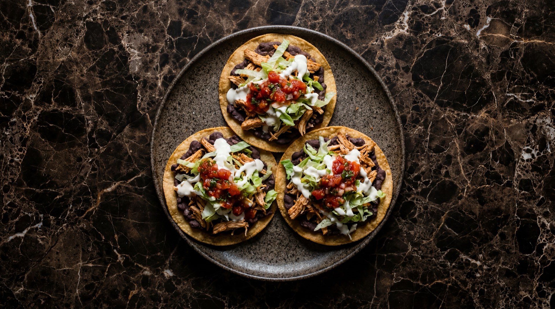 Tostadas mexicanas de tinga de pollo con crema acida y aguacate sobre plato de terracota oscura en mesa de marmol nero marquina con vetas blancas