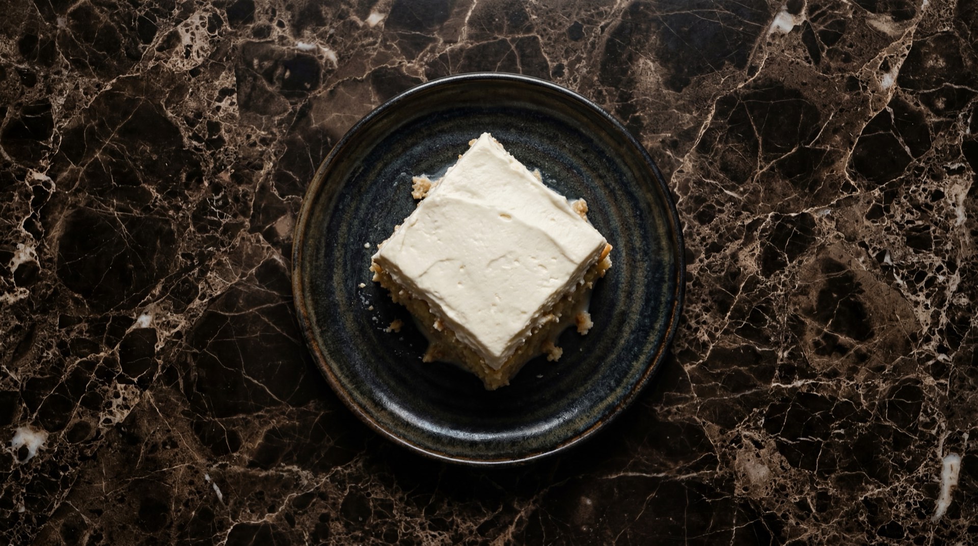 Pastel de tres leches mexicano con crema batida y canela en polvo sobre plato de ceramica oscura en mesa de marmol negro con vetas doradas