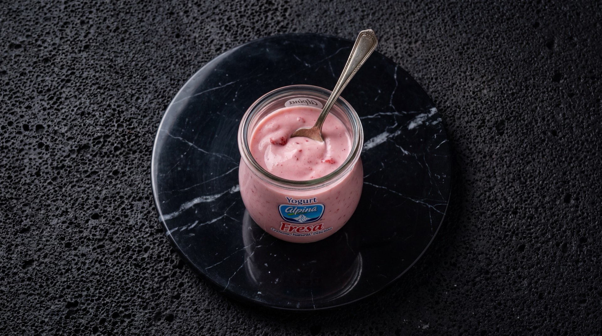 Yogurt Alpina natural en vaso de vidrio sobre mesa de marmol oscuro, textura cremosa blanca con cultivos lacticos vivos